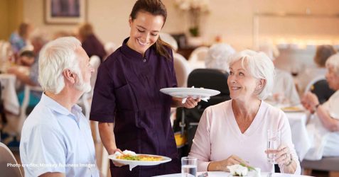 Older residents dining together in a bright, home-like aged-care dining room, with a staff member engaging with them warmly.